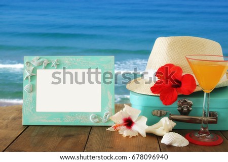 Fedora hat, sunglasses, tropical hibiscus flower next to blank frame over wooden table and beach landscape background. relaxation or vacation concept. For photography montage