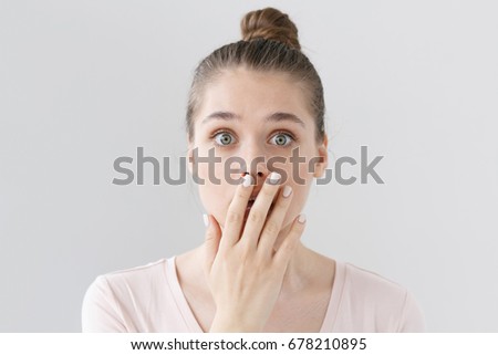 Indoor shot of beautiful teenage girl isolated on gray background dressed in light pink top, covering mouth with hand in sign of deep amazement and shock, hardly believing news she is receiving.