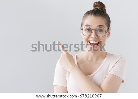 Horizontal picture of young good-looking female isolated on grey background wearing big round spectacles, pointing leftwards with forefinger, looking extremely happy, satisfied and surprised.