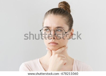 Headshot of good-looking young female in glasses isolated on gray background with head turned leftwards, scratching chin with fingers in deep thinking with uncertain expression and doubt on face. Royalty-Free Stock Photo #678211060