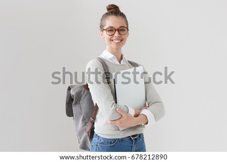 Indoor photo of college student girl isolated on gray background, smiling at camera, pressing laptop to chest, wearing backpack, ready to go to studies, start new project and suggest new ideas.
