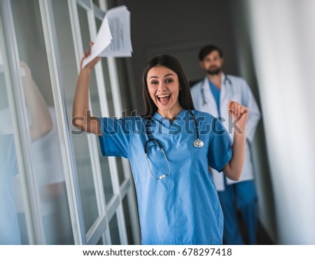 Beautiful young doctor in blue scrubs is holding paper and screaming with happiness while standing in hospital hall, her colleague in the background