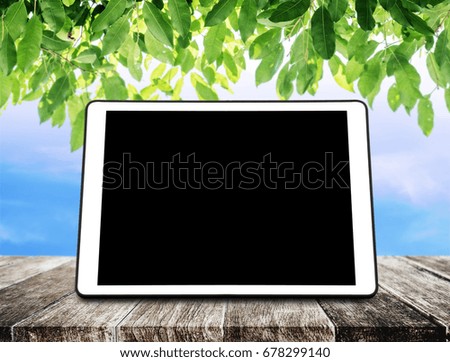 Digital tablet on wooden table with green leaves and blue sky background, with black empty screen