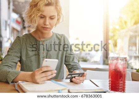 Elegant blonde woman in blouse sitting at cafe holding mobile phone and plastic card signing up on website. Businesswoman with stylish hairdo paying with credit card while shopping online using mobile