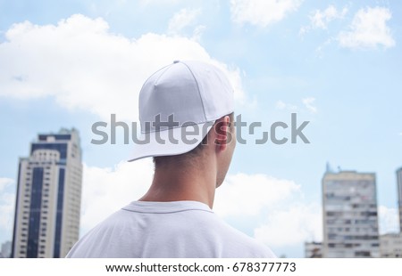 Young man in a white baseball cap and white t-shirt on background of the city and blue sky with clouds Royalty-Free Stock Photo #678377773