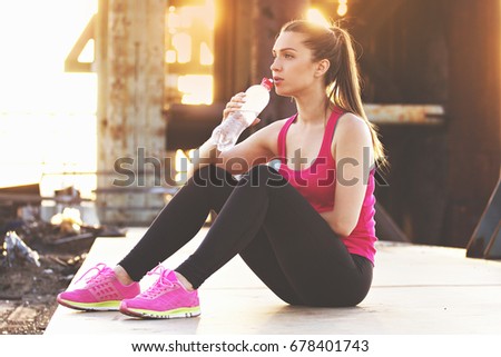 Staying hydrated. Beautiful young woman in sports clothing drinking water while having rest after workout. Evening sunlight and industrial view on background.