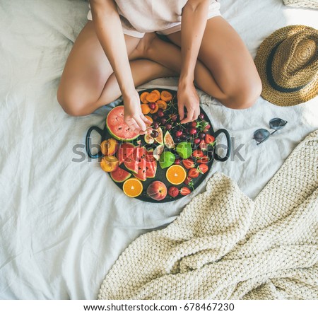 Summer healthy raw vegan clean eating breakfast in bed concept. Young girl wearing pastel colored home clothes taking cherries from tray full of fresh seasonal fruit. Top view, square crop, copy space Royalty-Free Stock Photo #678467230
