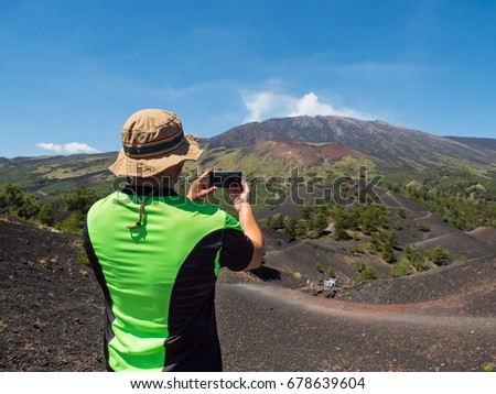 tourist photographer take a panoramic picture, with his smarphone, of the beautiful valley of Etna Vulcano with, craters, vegetation and lava stone during an excursion . satured color filter