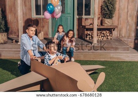 girls sitting on porch while boys playing with cardboard airplane
