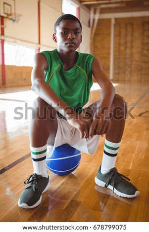 Portrait of teenage boy with hands clasped sitting on basketball in court