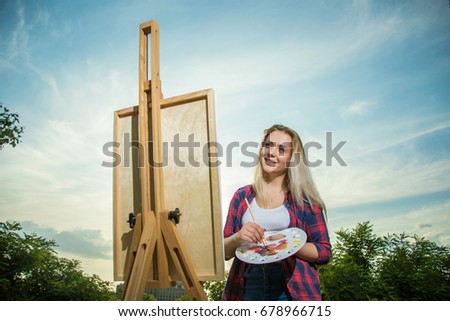 A woman artist with a brush and polytray in her hands draws on canvas against the backdrop of nature. The artist draws on the easel. Artist at work
