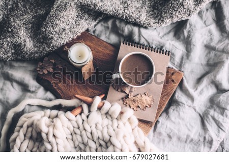 Cup of coffee and candle on rustic wooden serving tray in the cozy bed with blanket. Knitting warm woolen sweater in the autumn weekend, top view.