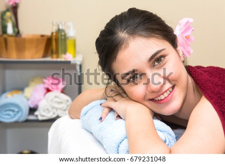 Portrait of young beautiful woman smiling and lying on the massage bed in spa salon with spa decoration. Happy attractive girl with tropical flower relaxing and looking at camera in spa resort.
