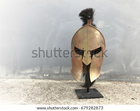 Gold lron Roman helmet placed in front of gray wall.