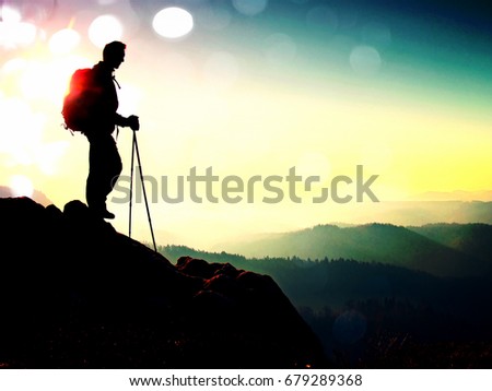 Film grain effect.  Tourist with sporty backpack and poles in hands stand on rocky view point and watching into deep misty valley bellow. Sunny spring daybreak in rocky mountains.
