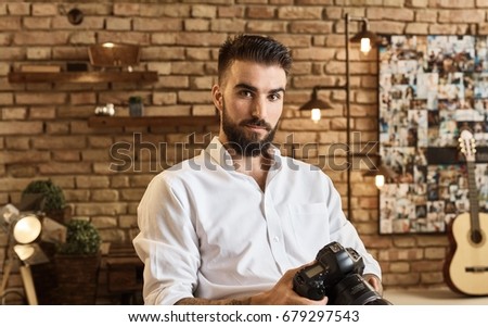 Portrait of bearded young man holding camera at trendy industrial loft home. Photographer portrait.