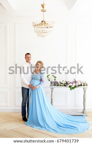 A young handsome man is hugging a beautiful pregnant girl. They put their hands on the girl's belly. They stand in a bright room, an aristocratic interior. She is dressed in a long blue dress. 