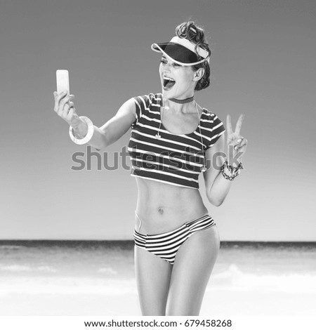Perfect summer. smiling young woman in red sun visor on the beach with cellphone taking selfie and showing victory gesture