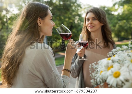 Picture of pretty young two women sitting outdoors in park drinking wine. Looking aside.