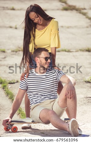 young man sitting on skateboard while his african american girlfriend standing behind