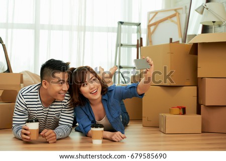Loving young couple taking selfie on mobile phone while lying on floor and drinking fragrant coffee from paper cups, pile of moving boxes and interior design items on background