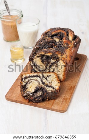 Sweet homemade poppy seeds braided (twisted) yeast bread with walnuts on a cutting board. Milk, cane sugar and honey in jars on a white wood background. Vertical orientation