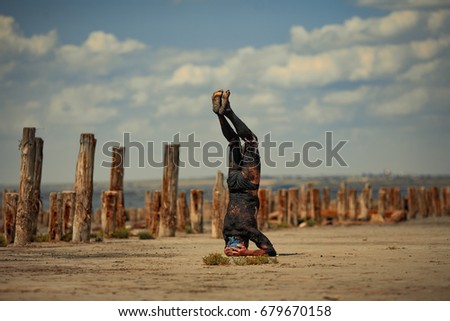 Young man smeared with therapeutic mud and stands on head on beach background. Next there are wooden columns. Spa.