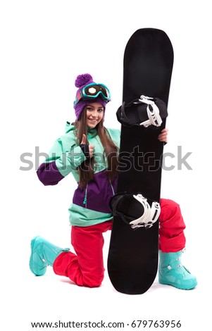 vertical picture, brunette pretty young caucasian woman in purple ski outfit, blue snow boots and blue ski goggles stand on one knee and hold black snowboard, look at camera, show thumbs up