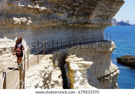 Female tourist walking on a passage carved into the cliffs near the sea level, below the medieval citadel of Bonifacio.