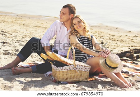 Loving couple. Horizontal shot of beautiful young woman looking to camera and smiling while sitting with boyfriend on beach and having picnic.