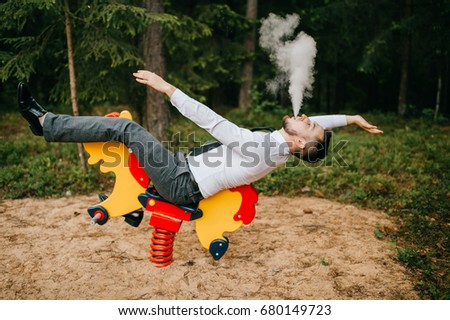 Adult serious man in business clothes riding children metal horse attraction with spring on playground. Odd person blows out clouds of thick smoke. Strange posing with arms apart. Weird behavior.