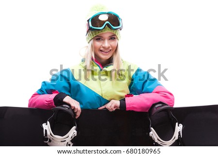 vertical picture, isolated on white, blonde pretty young caucasian woman in colorful ski outfit, yellow trousers and blue snow glasses sitting cross-legged behind black snowboard, look at camera