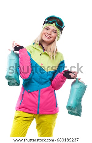 vertical picture, isolated on white, blonde attractive young caucasian woman in colorful coat and blue snow goggles, hold blue snow boots, look at camera