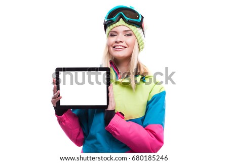 vertical picture, isolated on white, blonde beauty young caucasian woman in colorful ski suit and blue snow glasses hold black empty tablet