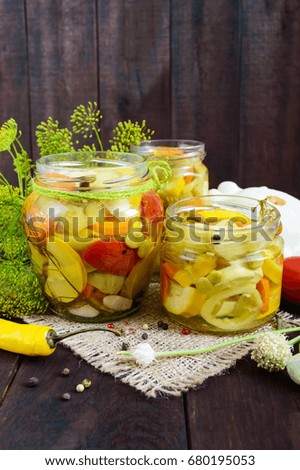 Pickles: vegetable assortment (zucchini, pepper, carrots, tomato, green peas) in glass jars on a dark wooden background.