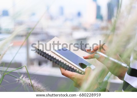 young woman using smart phone for work with notebook and pen at park of rooftop on city view.