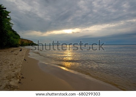 A serene picture of a sandy beach. The rising sun is coming out through the clouds and is beautifully reflected in the water. Trees in the background. 