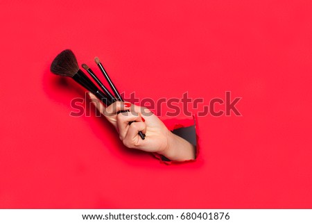Woman's hand holding out several make up brushes through crack in red wall.