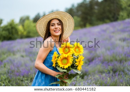 Beautiful young woman holding a bouquet of sun flower in the middle of a lavender field, wearing a elegant blue dress and a straw hat, looking at camera and smiling.