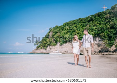 Beautiful couple having fun and walking along a white sandy beach with blue sky and turquois water on the background. Men and women are happy, they looking to each other and smiling together.