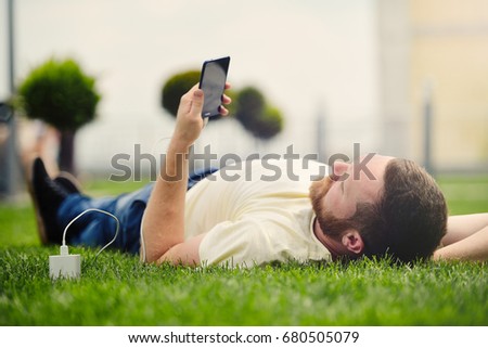 Nature and technology. A man with a beard lies on a green grass with a smartphone in his hands is charging power bank  Royalty-Free Stock Photo #680505079