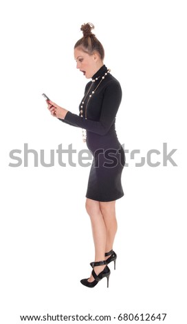 A shocked young woman in a black dress and long necklace looking
with an open mouth at her phone, isolated for white background.
