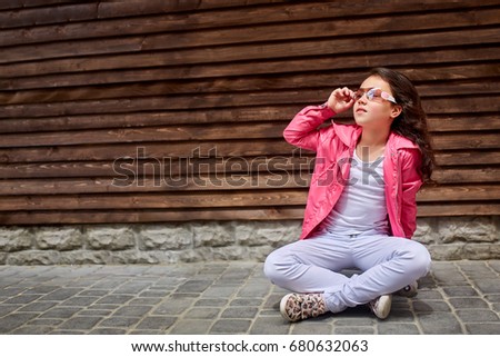 Fashion kid. Stylish little girl child wearing a summer or autumn pink jacket, white jeans, sunglasses with skateboard having fun in city over wooden wall on background