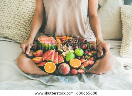 Summer healthy raw vegan clean eating breakfast in bed concept. Young girl wearing pastel colored home clothes sitting and holding tray full of fresh seasonal fruit Royalty-Free Stock Photo #680665888