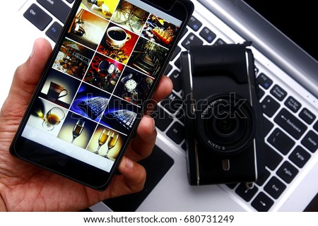 Photo of a hand holding a smartphone over a digital mirrorless camera and laptop computer