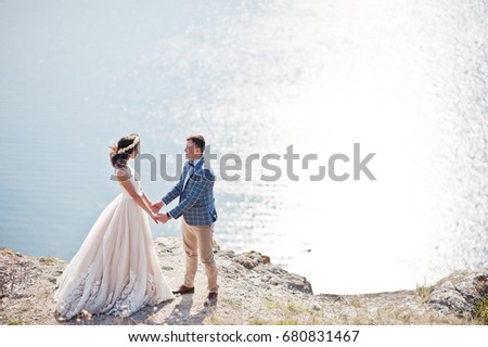 Fantastic wedding couple standing on the edge of rocky precipice with a perfect view of lake on the background.
