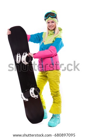 vertical picture, isolated on white, attractive  young caucasian girl in colorful ski costume, yellow trousers and blue ski glasses hold black snowboard, one leg on snowboard, look at camera