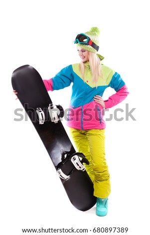 vertical picture, isolated on white, attractive  young caucasian girl in colorful ski costume, yellow trousers and blue ski glasses hold black snowboard, hand on waist, look at snowboard