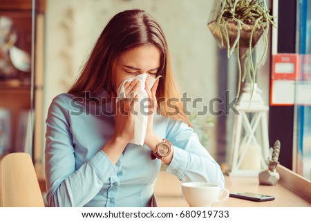 Flu. Young woman got nose allergy, flu sneezing nose sitting at the table in a trendy cafe coffee shop with a cup of hot beverage beside Royalty-Free Stock Photo #680919733