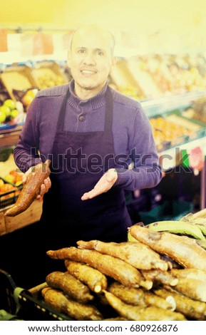 Professional seller posing near display of manihot roots in grocery and smiling